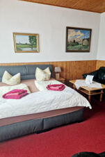 Jaukus pensiono "Zur Elbe" miegamasis su dvigule lova, raudonu kilimu, medinėmis dailylentėmis ir dviem paveikslais ant sienos.