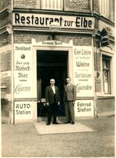 Istorinėje nespalvotoje nuotraukoje matyti restorano "Pension Zur Elbe" įėjimas su dviem vyrais prie&scaron;ais duris ir tipi&scaron;ka i&scaron;kaba.