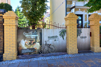 Įėjimas su vartais ir jaukių svečių kambarių reklama Pension Zur Elbe geltonai tinkuotame pastate su medžiais kieme.