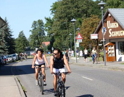 Zwei Personen auf Fahrr&auml;dern fahren eine sonnige Stra&szlig;e entlang, umgeben von B&auml;umen und kleinen Geb&auml;uden, Parkhotel Luckau-Cahnsdorf in der N&auml;he.