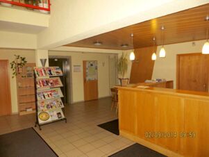 Reception area of Parkhotel Luckau-Cahnsdorf with wooden front desk, hanging lights, brochure stand, tiled floor, and beige walls