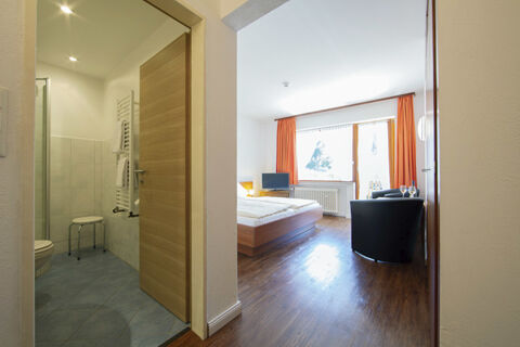 Světle zař&iacute;zen&yacute; dvoulůžkov&yacute; pokoj s dřevěnou podlahou, TV, oranžov&yacute;mi z&aacute;věsy a přilehlou koupelnou v Land-gut-Hotelu Heidsm&uuml;hle.