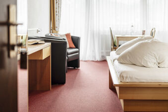 Helles Hotelzimmer im Hotel Engel mit rotem Teppich, Holzbetten, einem bequemen Sessel und einem kleinen Schreibtisch.