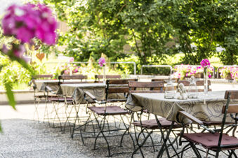 Sonnige Terrasse mit Tischen, St&uuml;hlen und Blumendekoration im Freien des Hotel Engel, umgeben von gr&uuml;nen B&auml;umen und Pflanzen.