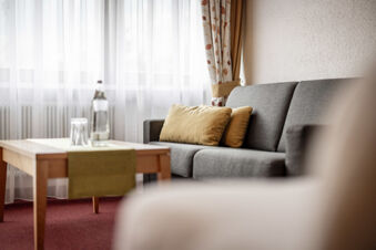 Gem&uuml;tliche Sitzecke mit grauem Sofa, gelben Kissen und Holztisch vor hellen Gardinen im Hotel Engel.