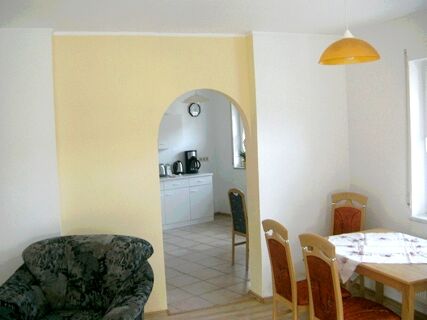 Living area with floral armchair, wooden dining table with four chairs, and kitchen visible through an arch in Fewos Privatpension Geib