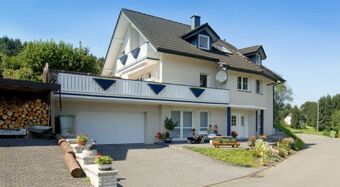 White multi-story house with balcony, garden, and driveway with two people sitting outside at Fewos Privatpension Geib