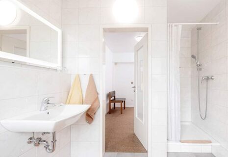 Helles Badezimmer im Hotel Dieck mit wei&szlig;em Waschbecken, zwei Handt&uuml;chern, Duschkabine und Blick in den angrenzenden Raum mit Stuhl.