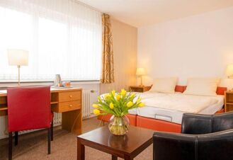 Helles Hotelzimmer im Hotel Dieck mit Doppelbett, Schreibtisch, Sitzbereich und einem Tisch mit einer Vase gelber Tulpen.