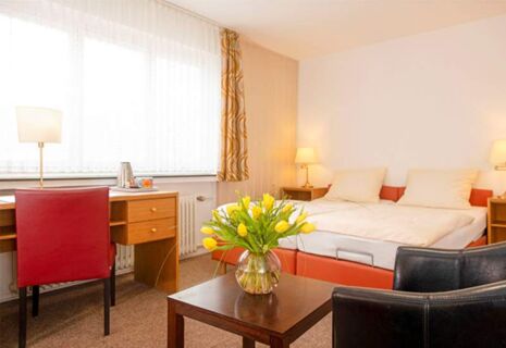 Helles Hotelzimmer im Hotel Dieck mit Doppelbett, Schreibtisch, Sitzbereich und einem Tisch mit einer Vase gelber Tulpen.