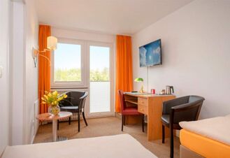 Helles und gem&uuml;tliches Hotelzimmer im Hotel Dieck mit wei&szlig;er Wand, orangenen Vorh&auml;ngen, Schreibtisch und Sitzbereich mit St&uuml;hlen und Tisch.