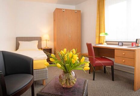 Helles Einzelzimmer im Hotel Dieck mit gelber Bettdecke, Holzschrank, Schreibtisch und einem Strau&szlig; gelber Tulpen auf dem Tisch.