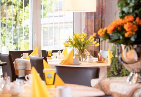Gedeckte Tische mit gelben Servietten und Blumenarrangements in einem hellen Speiseraum des Hotel Dieck, ideal f&uuml;r Fr&uuml;hst&uuml;ck.