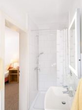 Helles Badezimmer mit Dusche, Waschbecken und beleuchtetem Spiegel im Hotel Dieck, Blick in angrenzendes G&auml;stezimmer mit Nachtischlampe.
