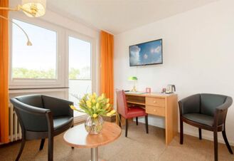 Helles und gem&uuml;tliches Zimmer im Hotel Dieck mit zwei Sesseln, Schreibtisch, Flachbildfernseher und gelben Vorh&auml;ngen.