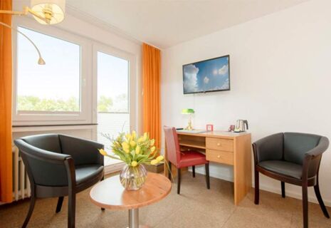 Helles und gem&uuml;tliches Zimmer im Hotel Dieck mit zwei Sesseln, Schreibtisch, Flachbildfernseher und gelben Vorh&auml;ngen.