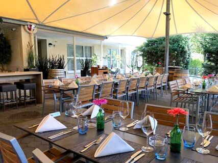 Au&szlig;enreich gedeckter Tischbereich unter Sonnenschutz mit Holzm&ouml;beln und Blumen im Hotel-Restaurant Felmis Nichtraucherhaus.