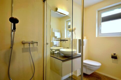 Shower area, glass partition, round sink with mirror light, hairdryer, wall-mounted toilet, and window inside Hotel-Restaurant Felmis Nichtraucherhaus