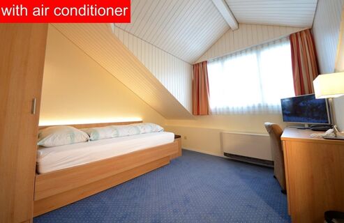 Single bedroom with slanted ceiling, wooden bed and furniture, blue carpet, window with curtains, TV and air conditioner at Hotel-Restaurant Felmis Nichtraucherhaus