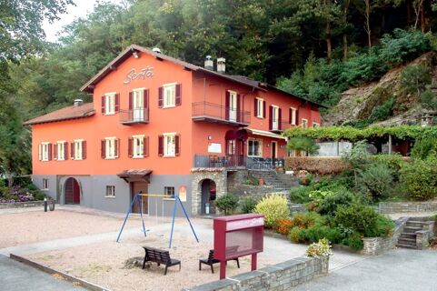 Vista exterior do edif&iacute;cio vermelho do Ristorante Albergo Grotto Serta com um parque infantil e uma colina verde ao fundo.