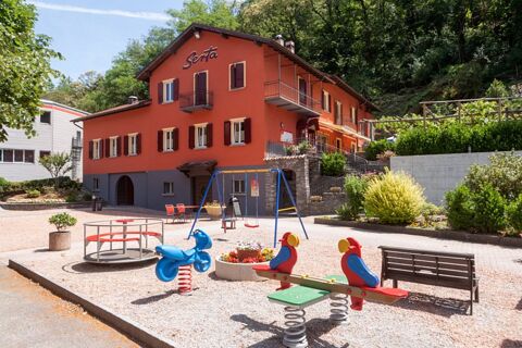 Playground equipment including a seesaw, spring rider, and swings in front of the red building of Ristorante Albergo Grotto Serta