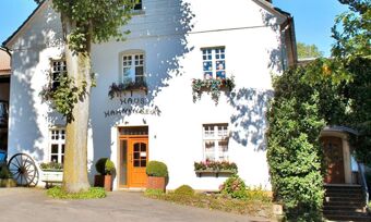 Die wei&szlig;e Fassade von Hotel Pension Haus Hahnenbecke mit Blumenk&auml;sten an den Fenstern und einer Holz-Eingangst&uuml;r an einem sonnigen Tag.