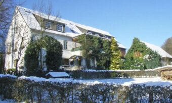 Das verschneite Geb&auml;ude von Hotel Pension Haus Hahnenbecke umgeben von B&auml;umen und einem Garten unter blauem Himmel im Winter.