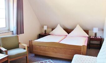 Helles und gem&uuml;tliches G&auml;stezimmer im Hotel Pension Haus Hahnenbecke mit Doppelbett, Sesseln und Nachttischen unter einem Dachschr&auml;genfenster.