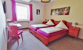 Hell eingerichtetes Doppelzimmer mit roten M&ouml;beln, violetten Bettauflagen und Fenster mit Vorh&auml;ngen im Hotel Pension Haus Hahnenbecke.