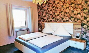 Modern eingerichtetes Schlafzimmer mit Doppelbett und floraler Tapete im Hotel Pension Haus Hahnenbecke, helles Fenster mit Vorh&auml;ngen und Nachttischlampe.