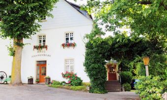 Au&szlig;enansicht des wei&szlig;en Geb&auml;udes mit Blumen an den Fenstern und begr&uuml;ntem Bogen am Eingang des Hotel Pension Haus Hahnenbecke.