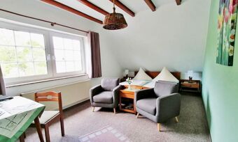 Helles Zimmer im Hotel Pension Haus Hahnenbecke mit zwei grauen Sesseln, Doppelbett, Holzm&ouml;beln und gro&szlig;em Fenster mit Blick ins Gr&uuml;ne.