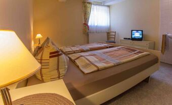 Gem&uuml;tliches Zimmer im Lindensch&auml;nke Hotel garni mit Doppelbett, Nachttischlampen, kleinem Fernseher und Fenster mit Vorh&auml;ngen.