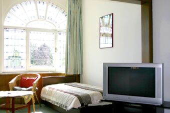 Ein helles Zimmer im Hotel-G&auml;stehaus Br&uuml;ggemann mit einem Einzelbett, Korbsessel, gro&szlig;em Fenster mit Vorh&auml;ngen und einem Fernseher.