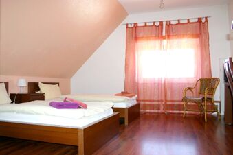 Helles Zimmer im Hotel-G&auml;stehaus Br&uuml;ggemann mit zwei Einzelbetten, Holzm&ouml;beln, roter Gardine und einem Korbstuhl am Fenster.