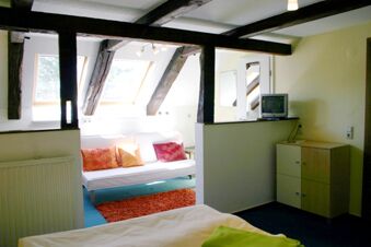 Hell eingerichtetes G&auml;stezimmer mit wei&szlig;en W&auml;nden und Holzbalken, ausgestattet mit Sofa, bunten Kissen und TV im Hotel-G&auml;stehaus Br&uuml;ggemann.