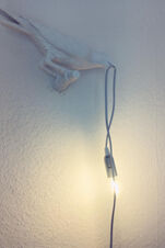 Eine Wandlampe in Form eines Vogels, der eine leuchtende Gl&uuml;hbirne h&auml;lt, dekorativ im Landhotel Alte M&uuml;hle installiert.