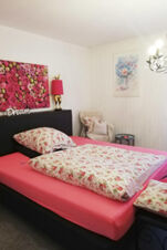 Helles Schlafzimmer im Landhotel Alte M&uuml;hle mit floralem Bettbezug, rosa Bettlaken, einem Sessel und dekorativen Bildern an der wei&szlig;en Wand.