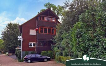 Au&szlig;enansicht des Landhotel Alte M&uuml;hle mit braunem Fachwerkgeb&auml;ude, vielen gr&uuml;nen B&auml;umen und geparktem Auto in einer ruhigen Stra&szlig;e.
