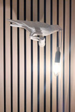 Eine Wandlampe in Form eines Vogels, der eine Gl&uuml;hbirne h&auml;lt, an einer Holzlattenwand im Landhotel Alte M&uuml;hle.