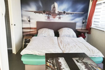 Gem&uuml;tliches Doppelzimmer im Landhotel Alte M&uuml;hle mit einem Bett, zwei Nachttischen und einem Wandbild eines landenden Flugzeugs.