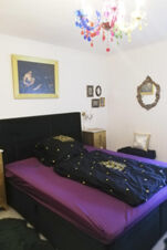 Schlafzimmer im Landhotel Alte M&uuml;hle mit einem lila bezogenen Bett, schwarzer Bettw&auml;sche, buntem Kronleuchter und dekorativen Bildern an der Wand.