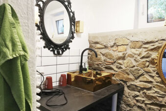Badezimmer im Landhotel Alte M&uuml;hle mit rustikaler Steinwand, goldenem Waschbecken, schwarzem Wasserhahn und dekorativem Spiegel.